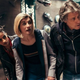 Doctor Who's Yaz in a leather jacket, the Doctor in her rainbow shirt, and Dan in a hoodie peer around a corner.